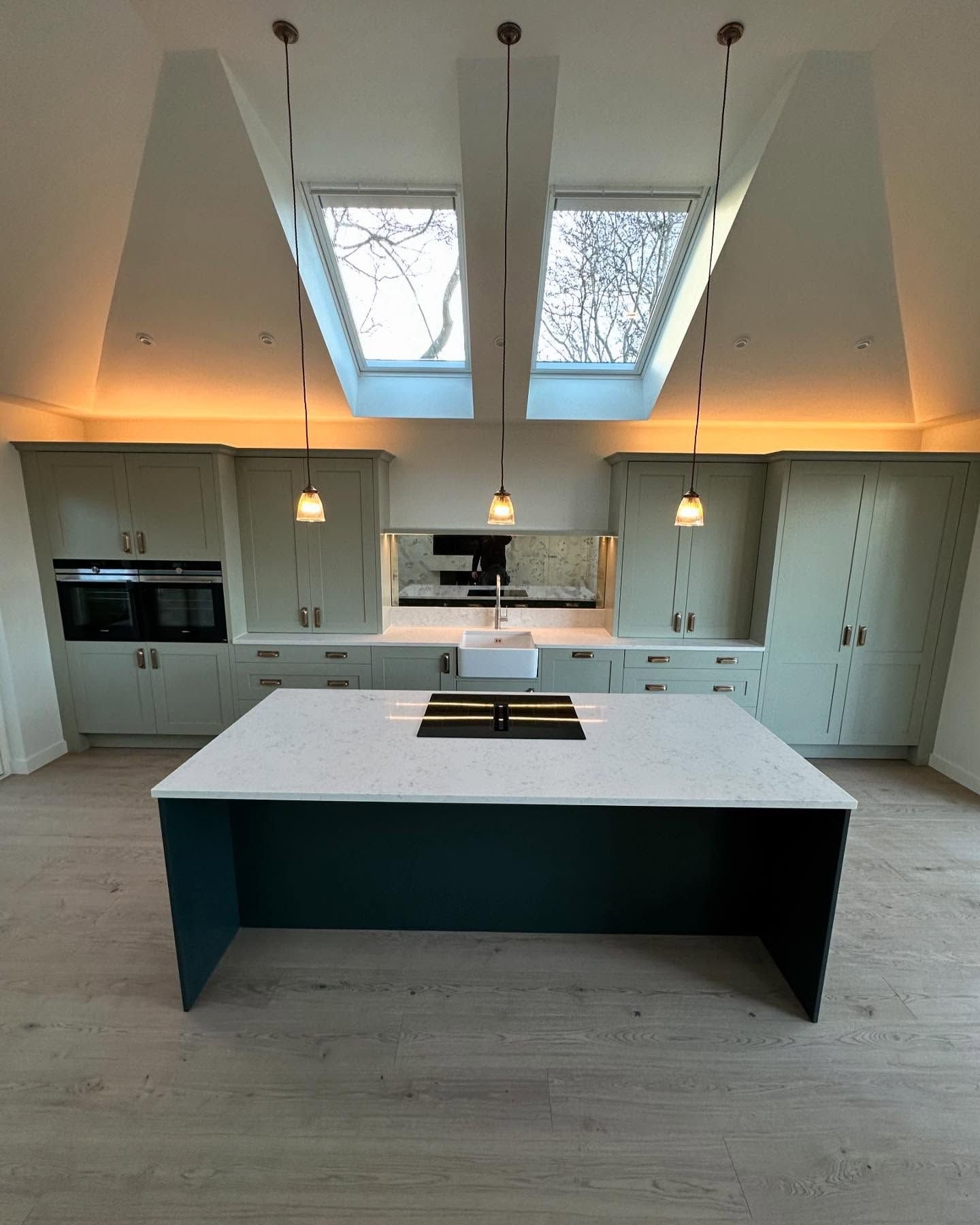 Kitchen in the converted golf shop: a vaulted ceiling with two roof windows, warm concealed lighting, sage hand-painted units and a dark island.