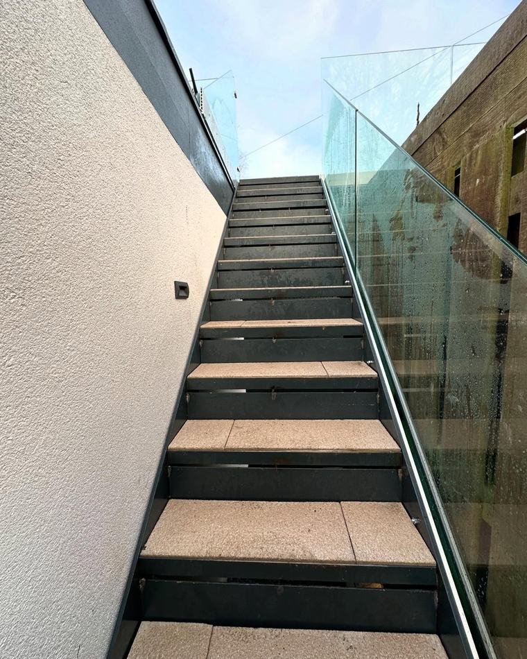 Steel stair with a frameless glass balustrade climbing along a rendered wall to the roof terrace.