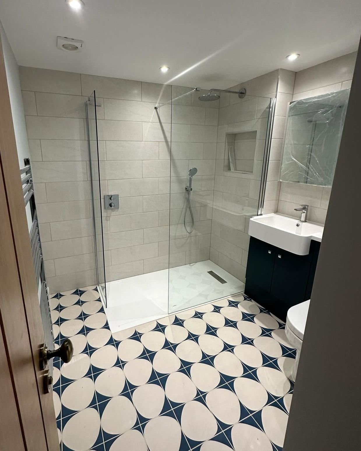 Finished basement shower room with a walk-in glass shower, pale wall tiles and a blue and white patterned floor.