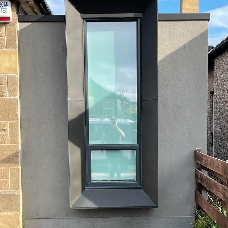 A tall window framed in black zinc, cantilevered out from a rendered side extension.